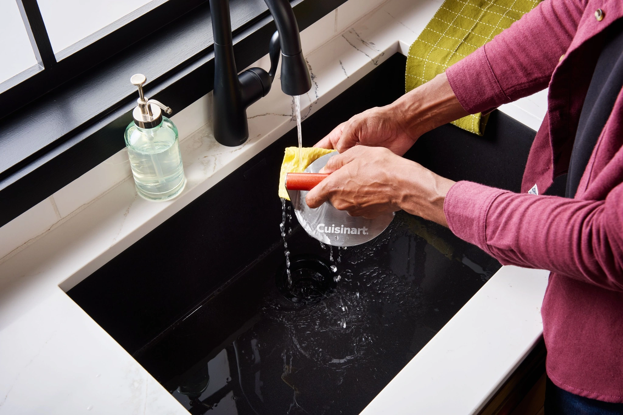 Hands washing a Cuisinart curved grilling tool under running water at a kitchen sink